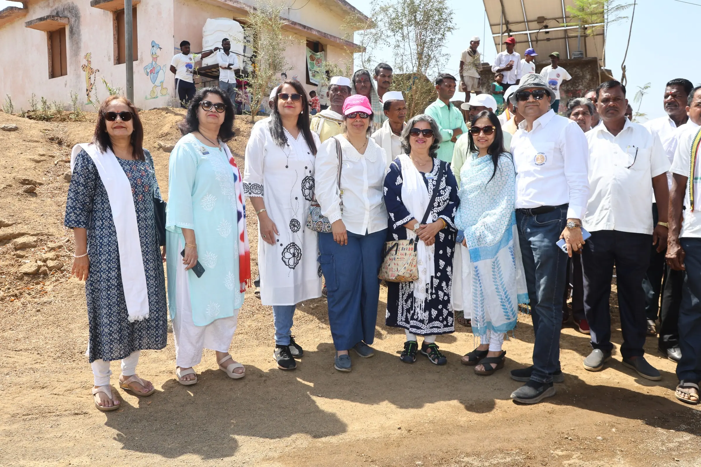 A group of women and men stand together on a dry hillside path in front of a village building