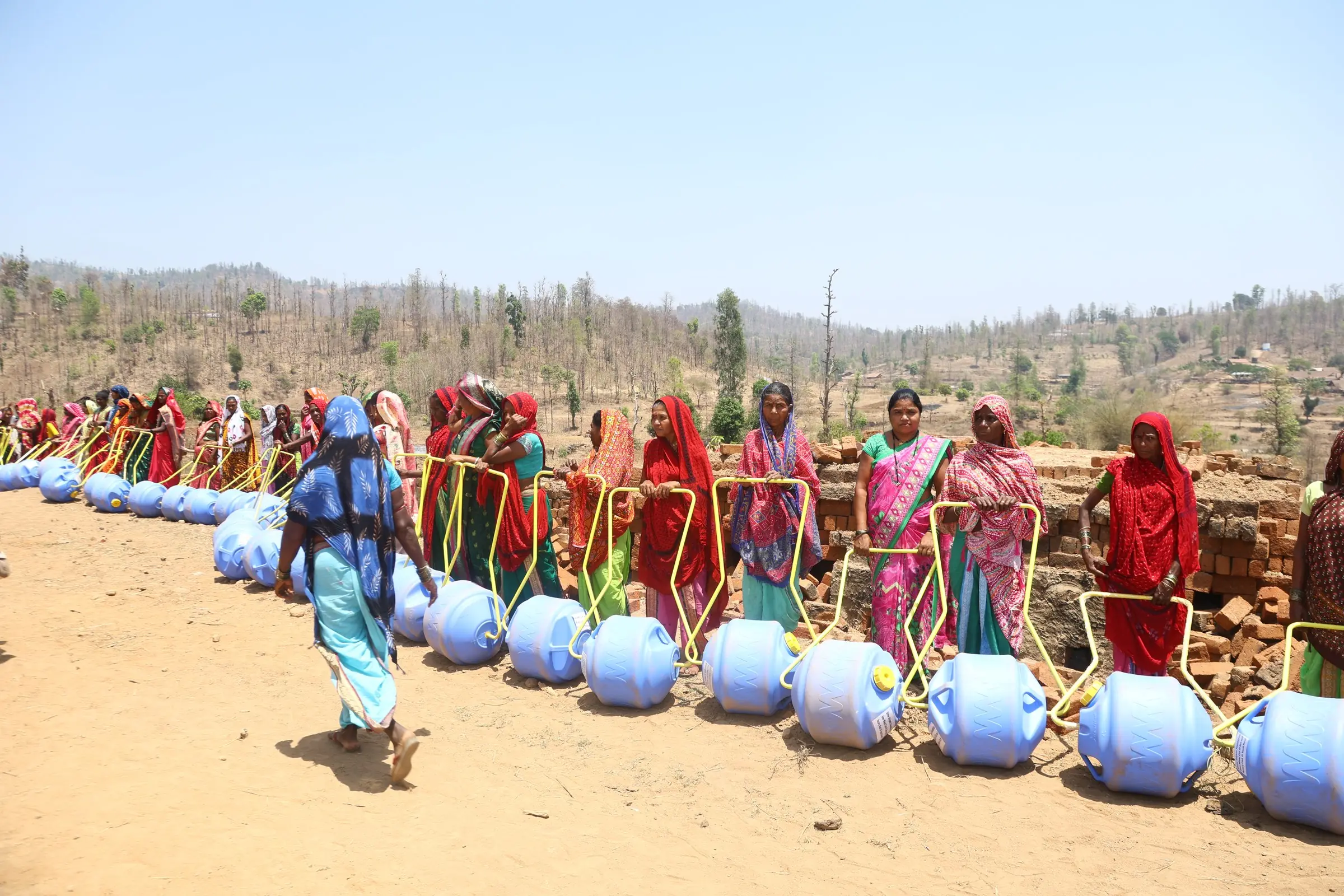 Women in red and pink saris stand in a line behind blue roller drums on dry ground, with hills behind