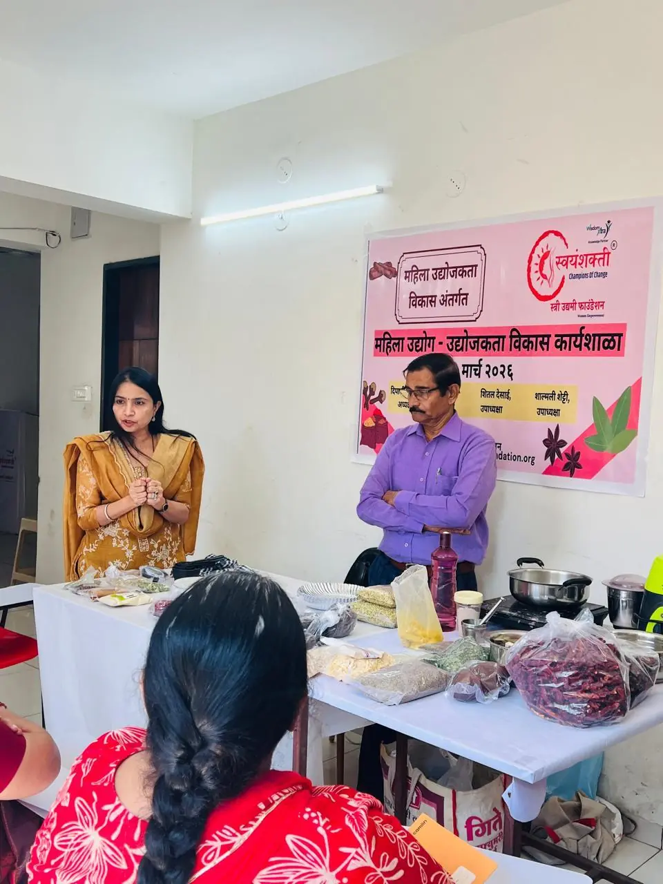 A woman and a man stand at a table of spices and cooking equipment in front of the workshop banner