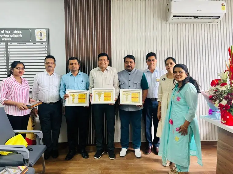 A group of people stand in a row in an office in front of a Marathi name board; three of them hold framed Doctors’ Day “Token of Gratitude” plaques