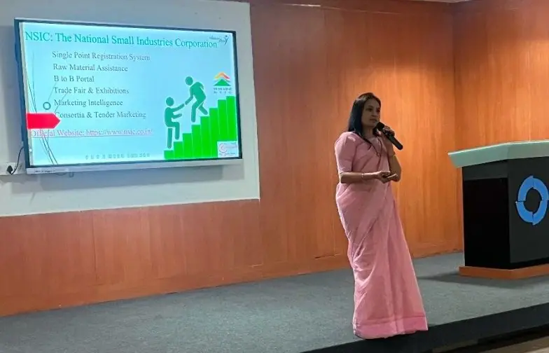 Woman speaking with a microphone beside a presentation slide about the National Small Industries Corporation